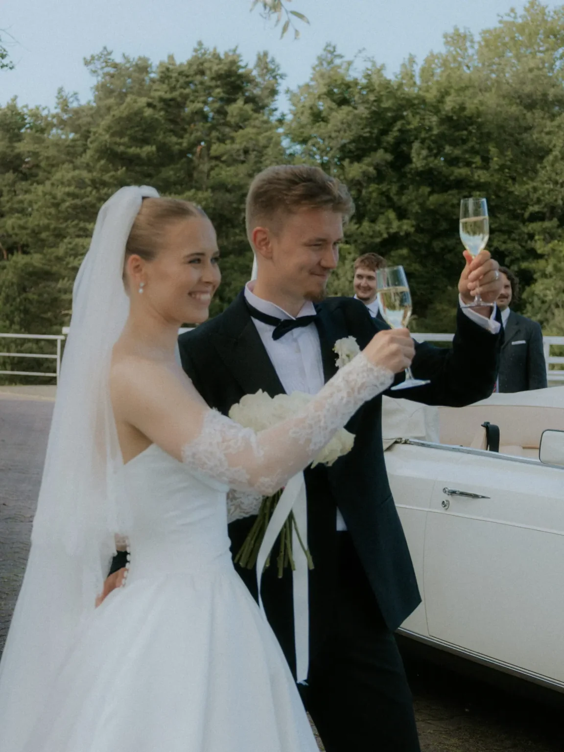 Hääpari kohottaa maljan valkoisen avoauton vieressä